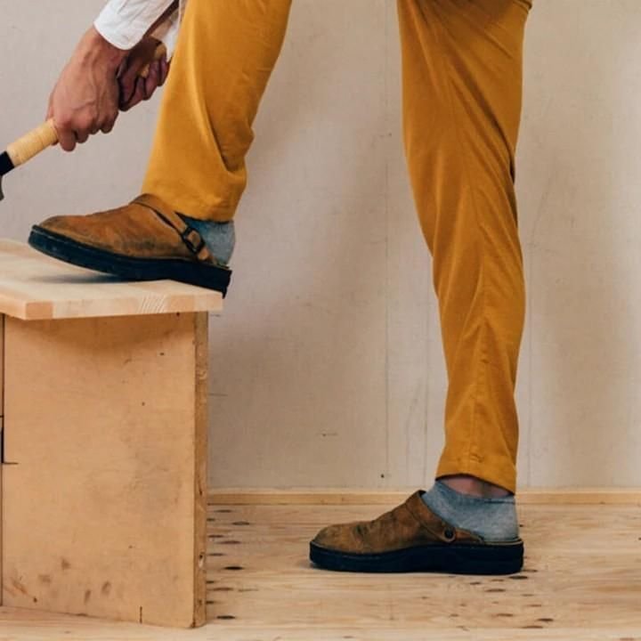Homme en pantalon jaune utilisant une scie sur une planche de bois, chaussures marron, intérieur minimaliste. Travail du bois, bricolage, artisanat.