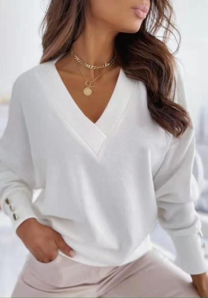 Femme portant un pull blanc à col en V, manches longues avec boutons, collier doré, posant élégamment. Mode féminine chic et décontractée.