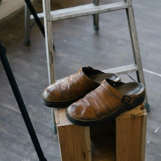 Sabots en cuir marron usés posés sur un bloc de bois, échelle en arrière-plan, sol en bois. Chaussures vintage, style rustique, intérieur atelier.