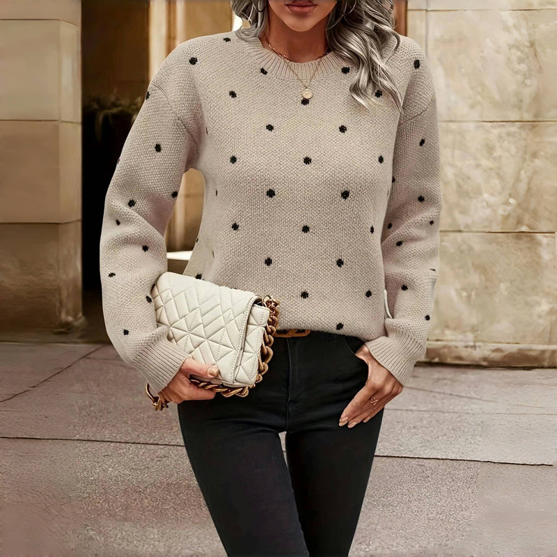 Femme portant un pull beige à pois noirs, tenant un sac à main matelassé blanc avec chaîne dorée, debout devant un mur en pierre beige.