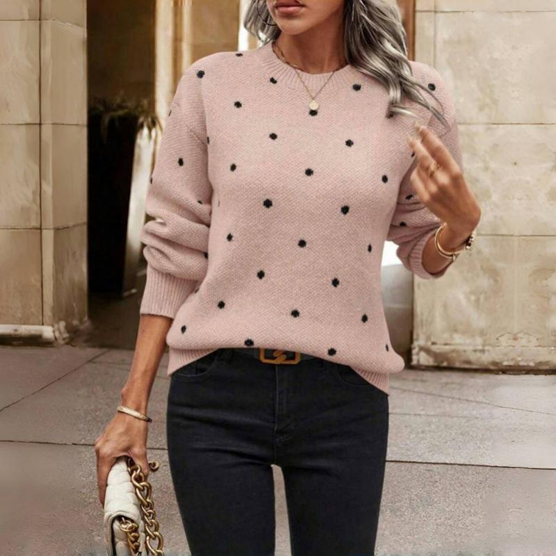 Femme portant un pull rose à pois noirs, jeans noirs, sac à main beige avec chaîne dorée, posant dans un cadre urbain élégant.