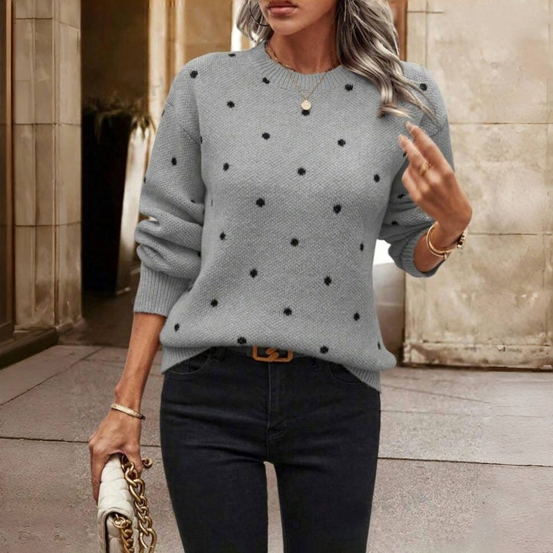 Femme portant un pull gris à pois noirs, manches longues, avec un sac à main blanc, dans un cadre urbain élégant. Mode automne chic.