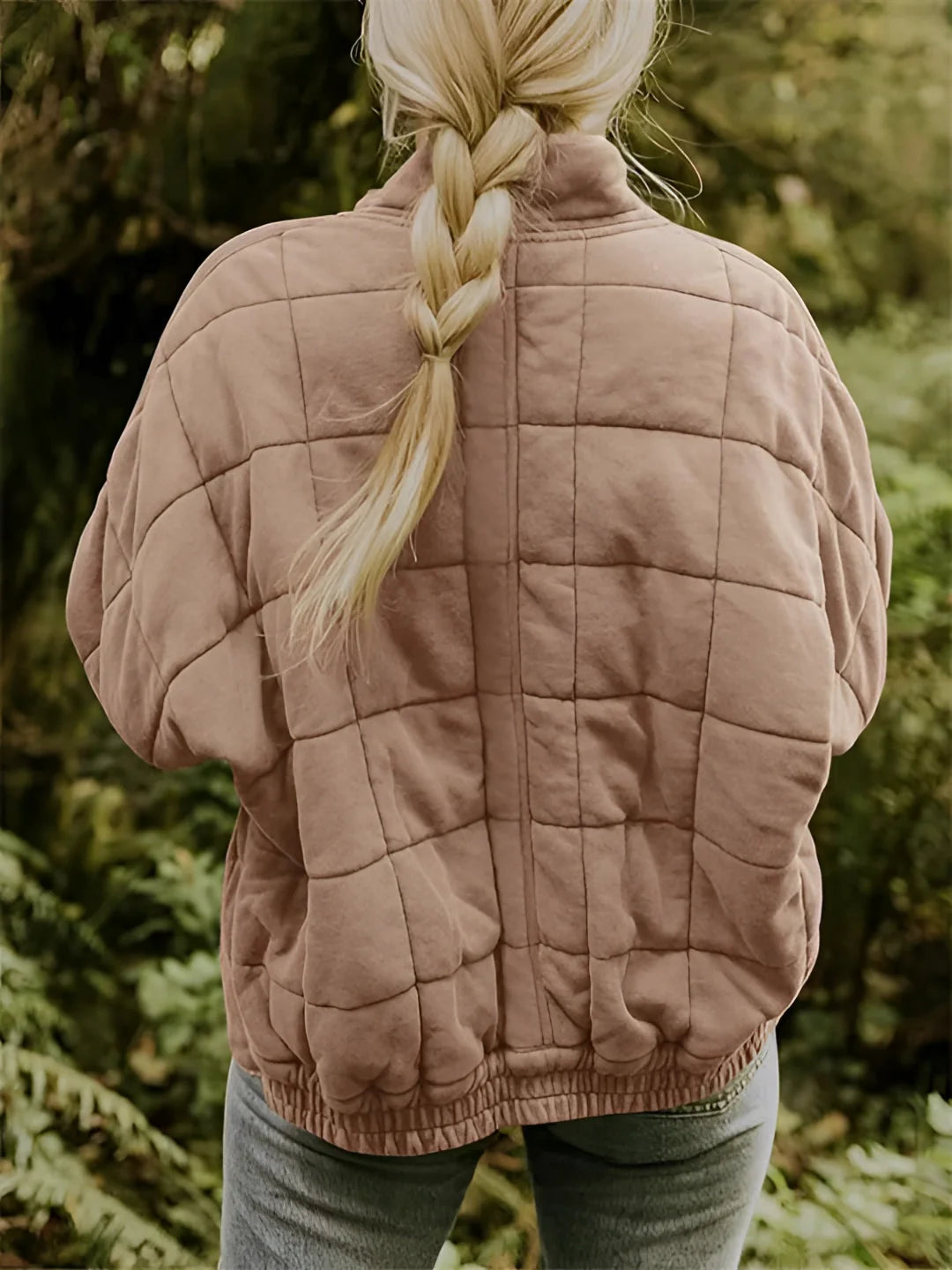 
A woman wearing a forest green quilted jacket stands in a natural setting, surrounded by grass and trees. The jacket features long sleeves and a lightweight, insulated design, with visible quilting patterns. The woman has long hair and holds a relaxed posture, showcasing the jacket's fit and style.