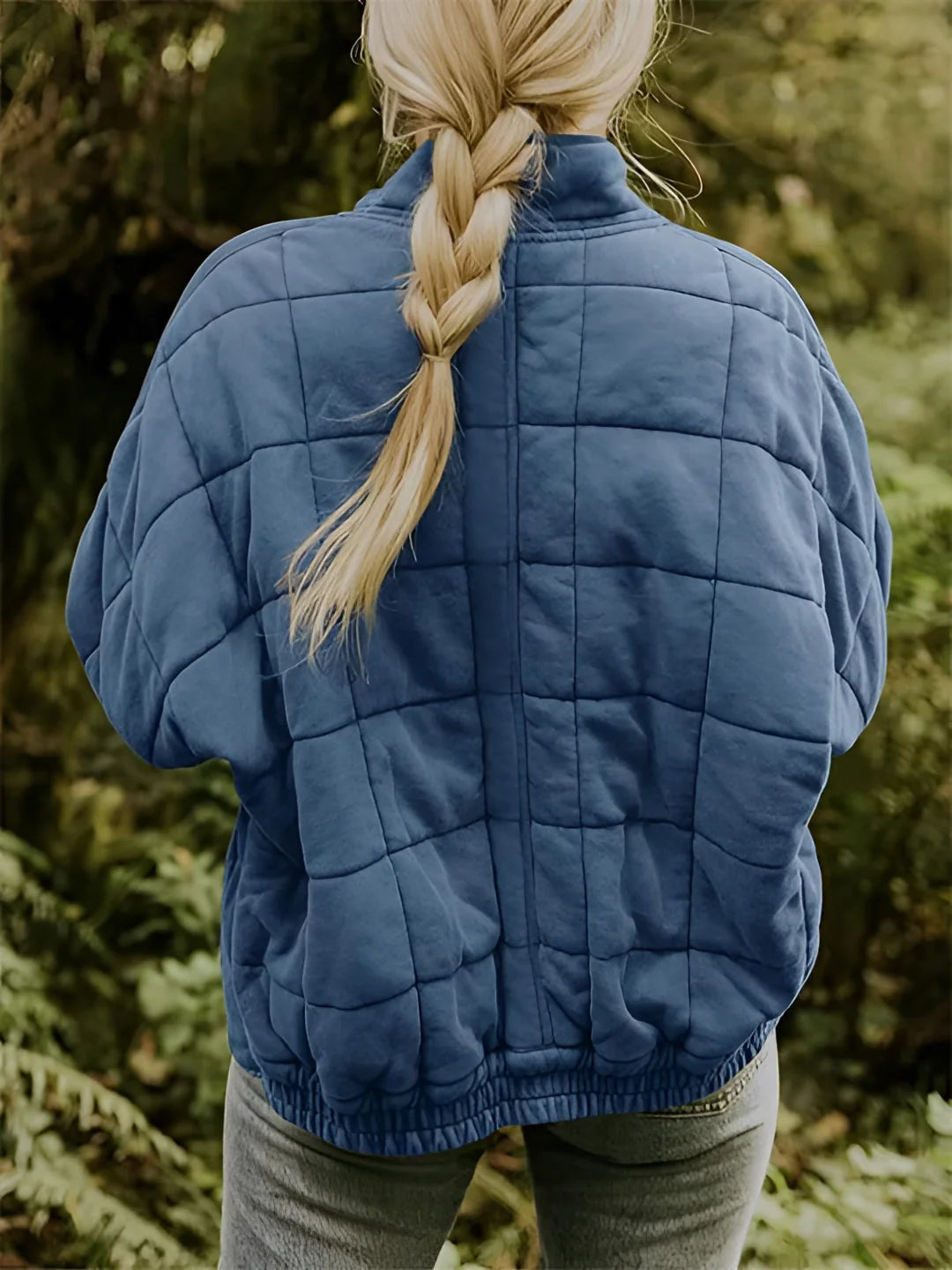 A woman wearing a forest green quilted jacket standing outdoors on grass. The jacket features lightweight insulation with a fitted waist and long sleeves. The woman pairs the jacket with a pair of grey denim jeans.