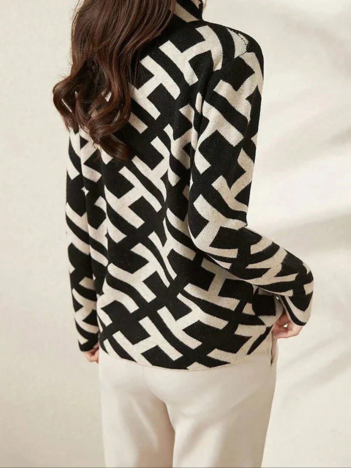 Femme portant un pull noir et blanc à motifs géométriques, dos tourné, cheveux bruns ondulés, pantalon beige. Mode automne-hiver tendance.