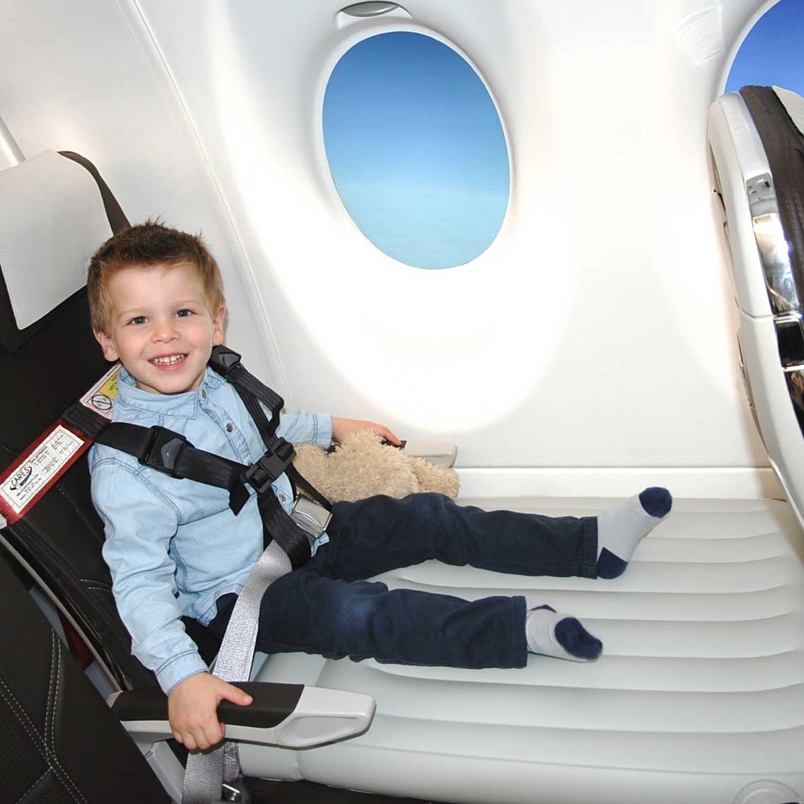 Lille dreng i flysæde med sikkerhedssele, smilende og holder en bamse, iført blå skjorte og jeans, ved siden af flyvindue med udsigt til blå himmel.