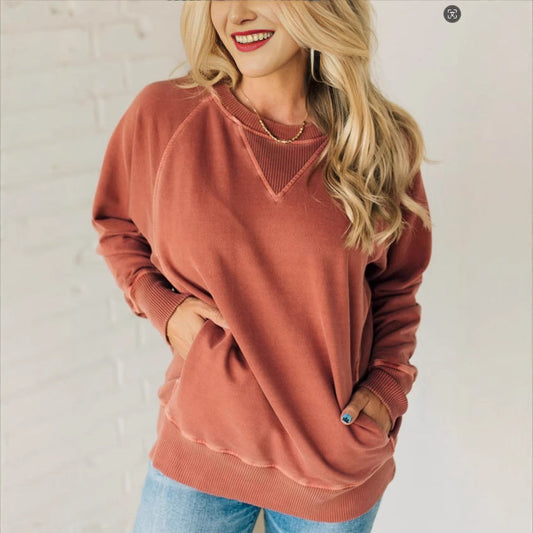 Femme souriante portant un pull rose saumon, manches longues, avec un collier doré, posant contre un mur blanc. Mode décontractée, style automne.
