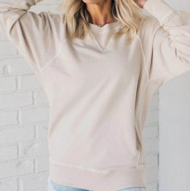 Femme portant un pull beige en coton, manches longues, devant un mur en briques blanches. Mode décontractée, style automne-hiver, tendance féminine.