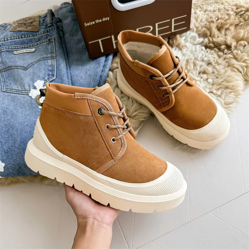 Brune ruskindsstøvler med hvid sål, placeret på beige gulv ved siden af blå jeans og en brun kasse. Moderne fodtøj, casual stil, komfortable sko.