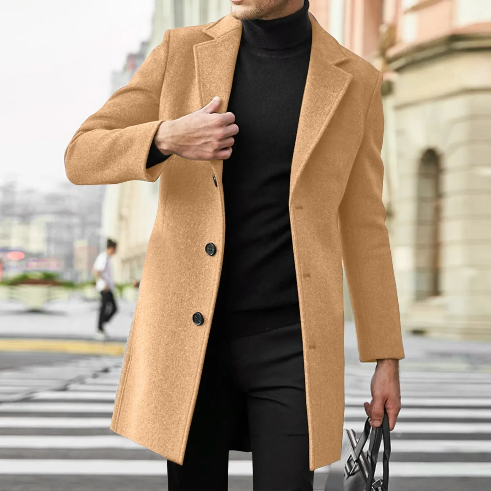 Mand i beige frakke krydser gade, bærer sort turtleneck og bukser, holder taske. Elegant vintermode, bymiljø, stilfuldt herretøj.