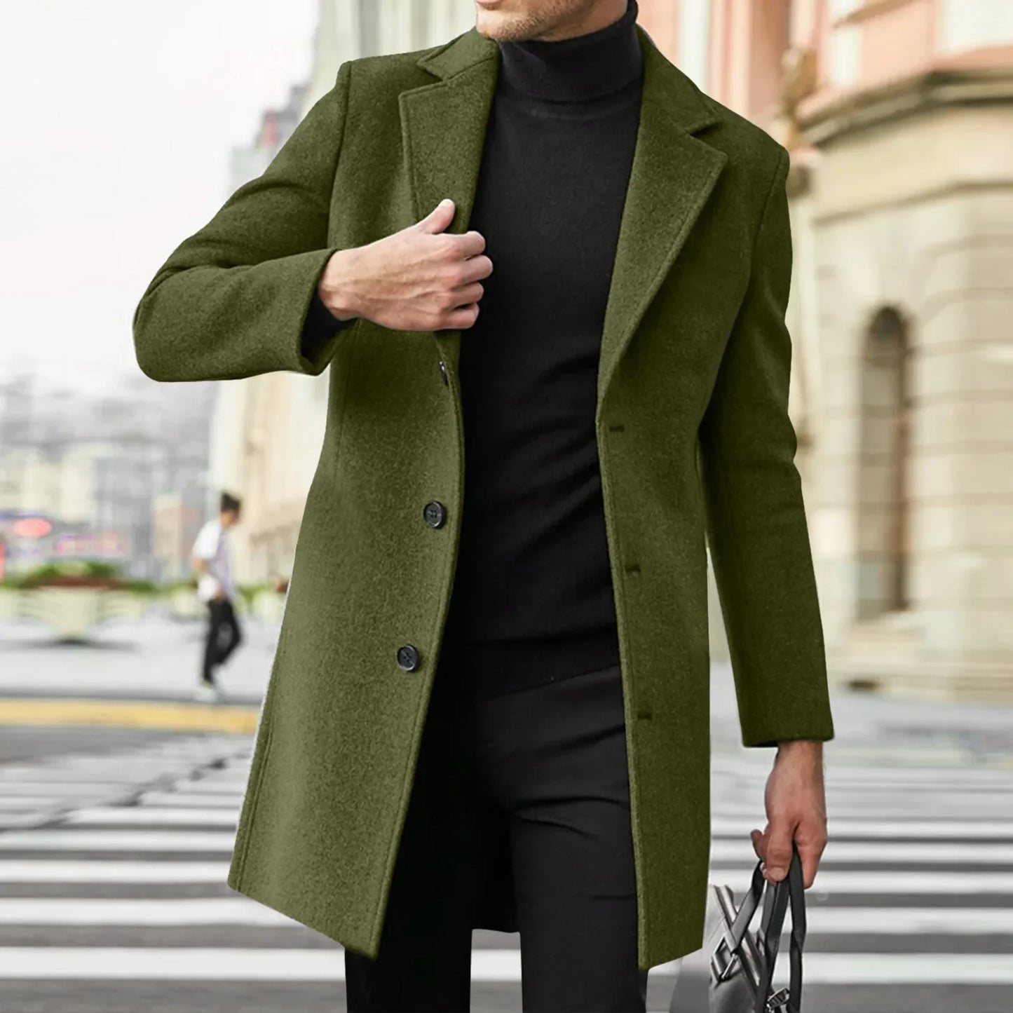 Mand iført olivengrøn frakke og sort turtleneck, krydser gade med bygninger i baggrunden. Stilfuldt herretøj, moderne mode, byliv.
