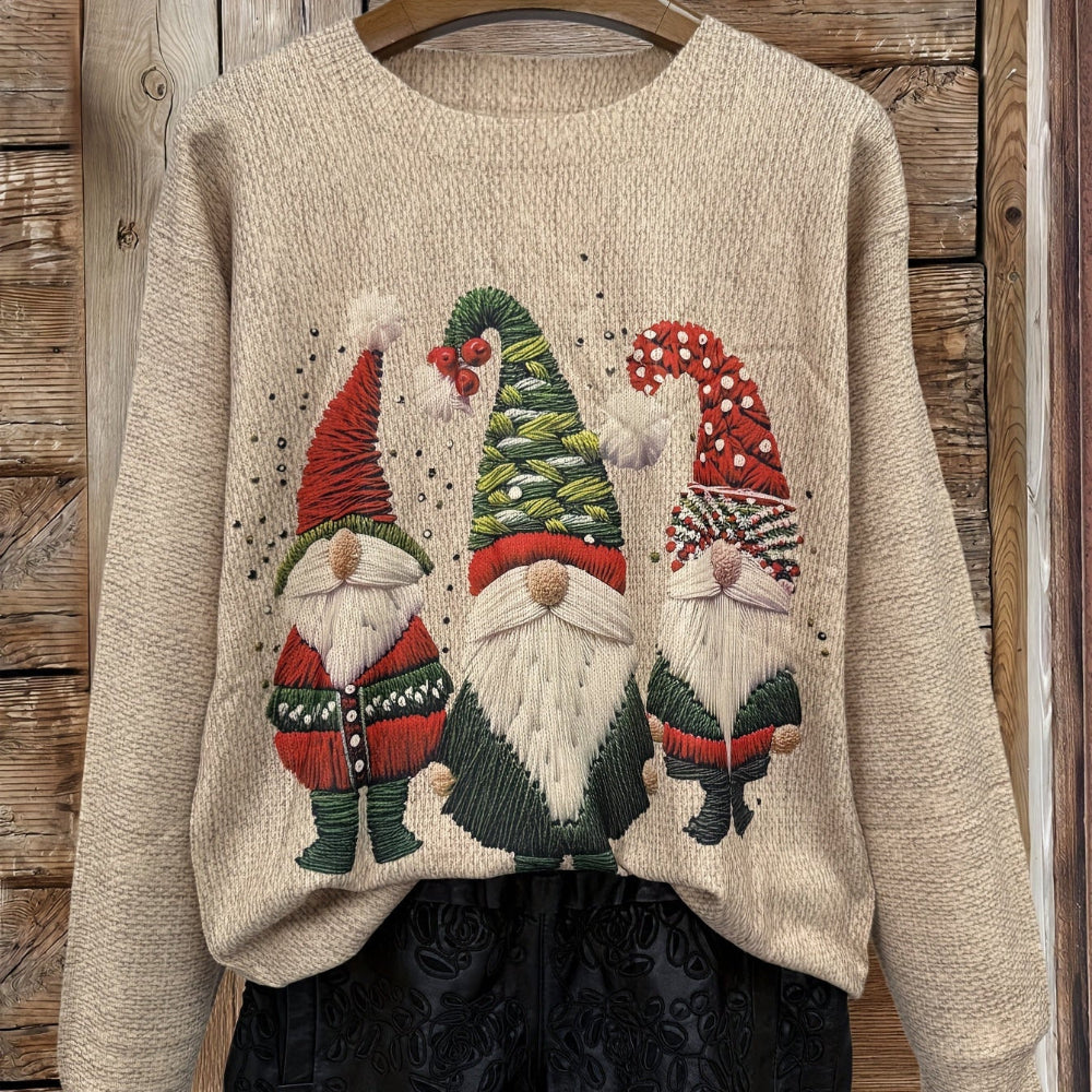 Pull de Noël beige avec motif de trois gnomes festifs, portant des chapeaux colorés, suspendu sur un cintre en bois devant une porte en bois rustique.