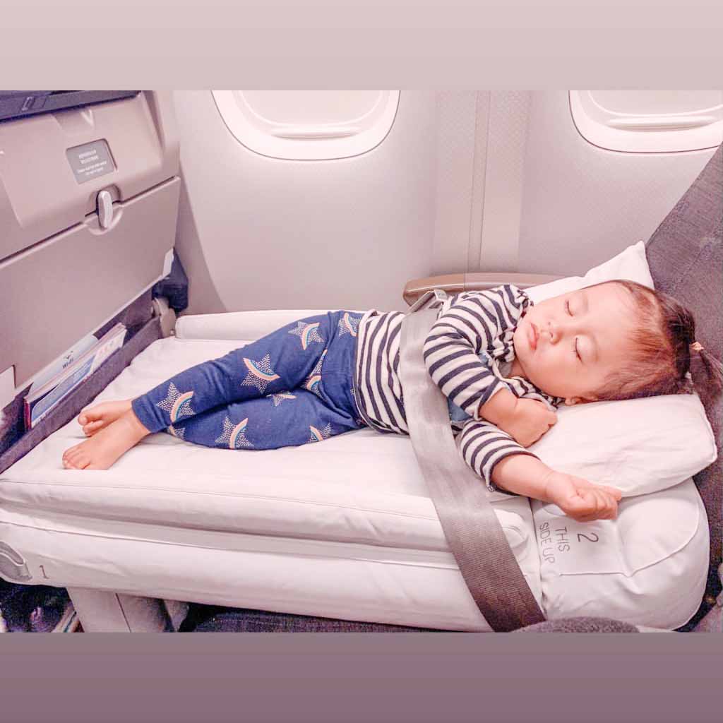 Lille barn sover komfortabelt på flysæde med sikkerhedssele, iført stribet trøje og stjernepyntede bukser. Rejse med børn, flykomfort.