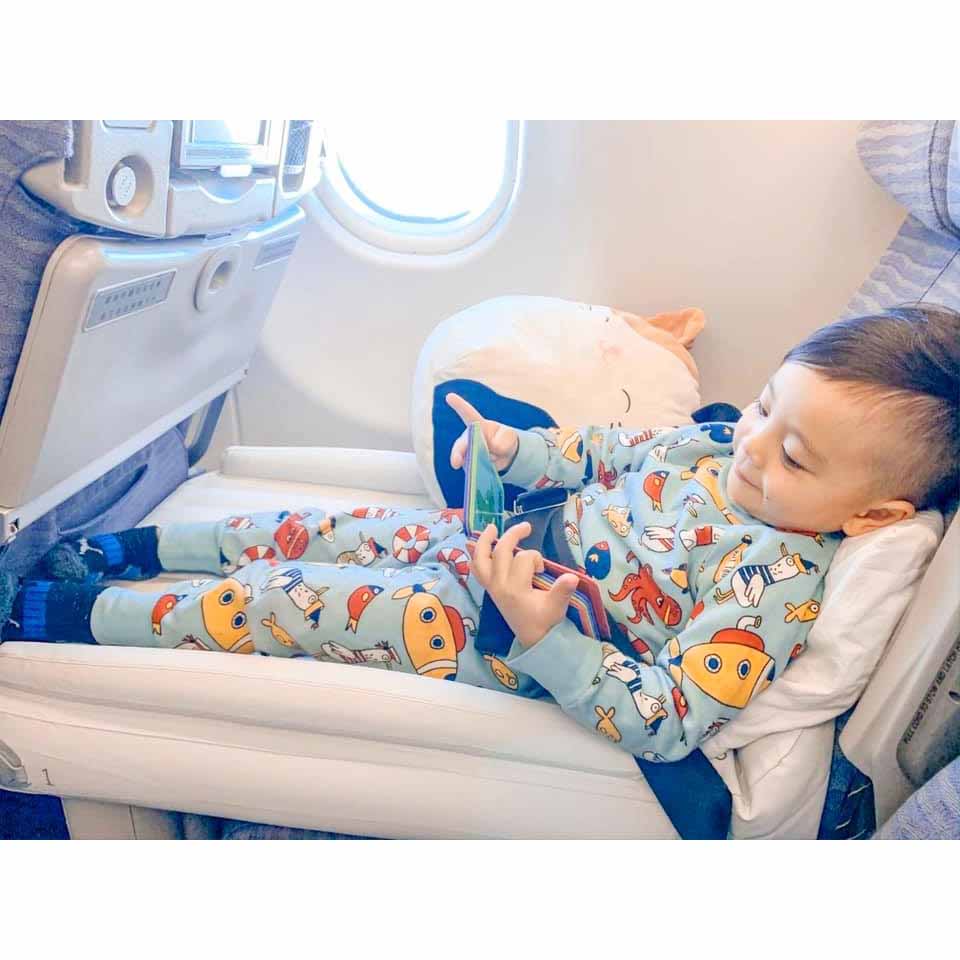 Lille barn i farverigt pyjamas sæt ser på smartphone i flysæde, rejser med fly, underholdning ombord, komfortabel flyrejse for børn.