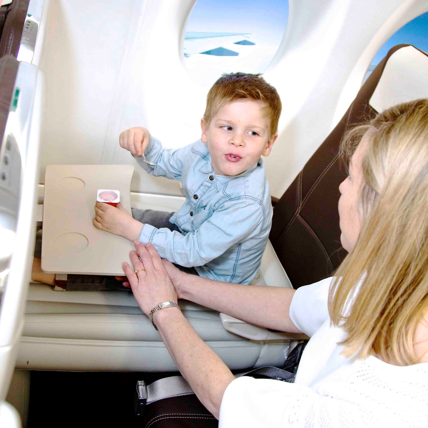 Dreng i flysæde med is, sidder ved siden af kvinde. Flyrejse, børnevenlig, komfortabel kabine, rejseoplevelse, familieferie, flyinteriør.